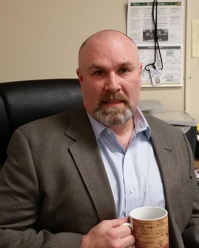 A man sits in a chair facing the camera. He has a goatee and is holding a coffee mug.