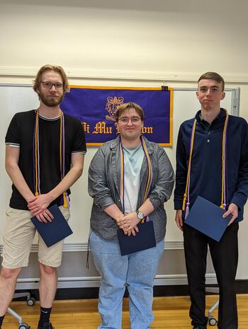 Three students stand in front of a Pi Mu Epsilon banner
