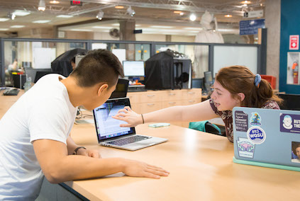 A picture of the CIT HelpDesk in Milne library.  A student is being assisted at the desk while 2 3D printers work in the background
