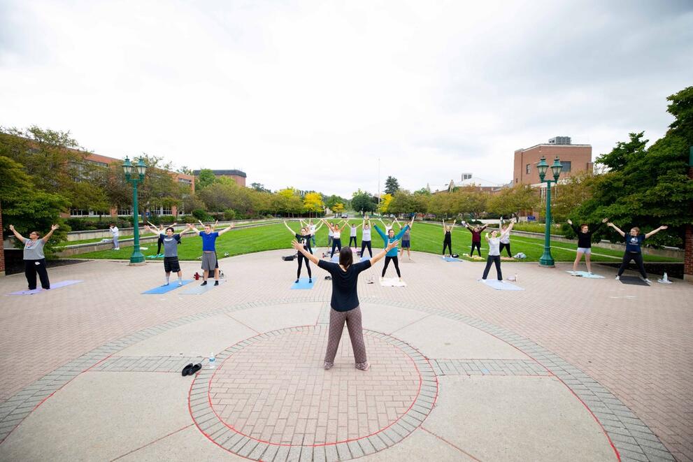 Beth Cholette with back to camera leading yoga class on brick area by College Green