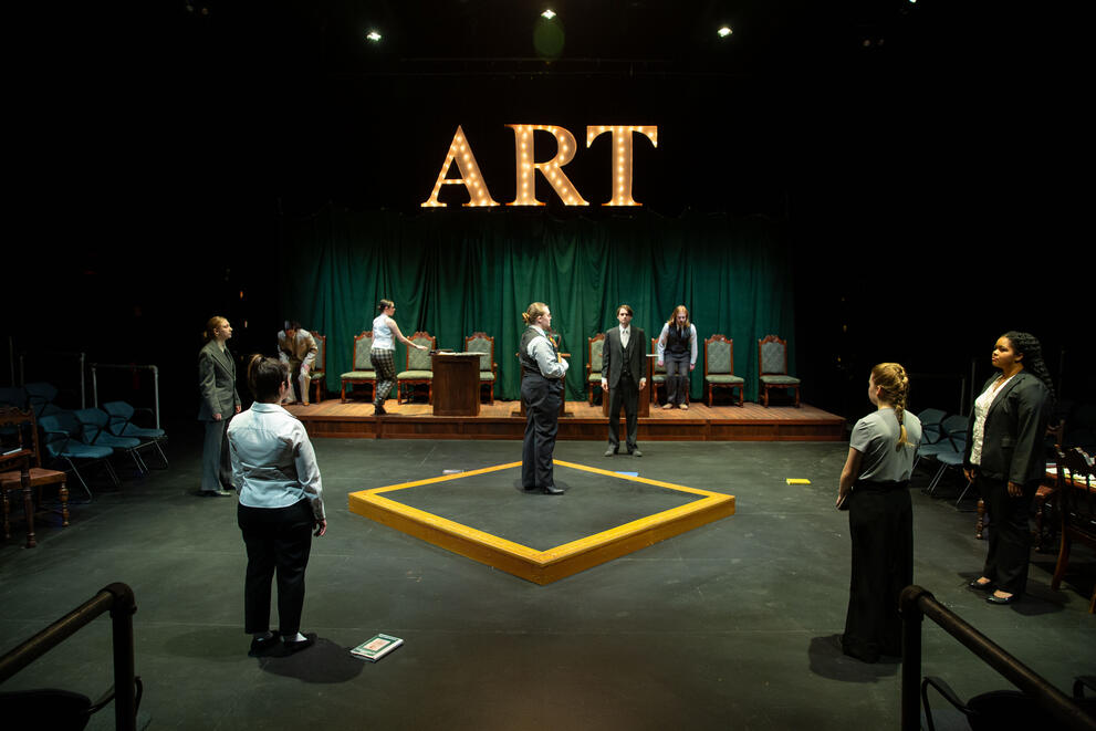 Stage setup with people standing around a yellow square on the floor. An illuminated sign reading 'ART' hangs above the stage. Green curtains and a row of chairs form the background.
