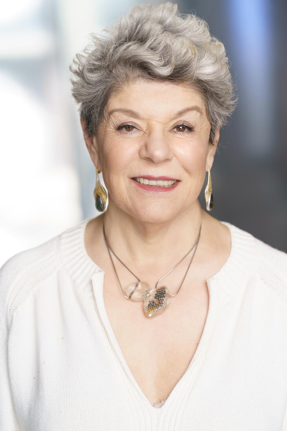 Image of Dr. Estela Bensimon with a smile wearing a cream v neck top.  Dr. Bensimon is wearing dangling earrings and a necklace with a large metal butterfly.  Dr. Estela Bensimon's hair is cut short and wavy. 