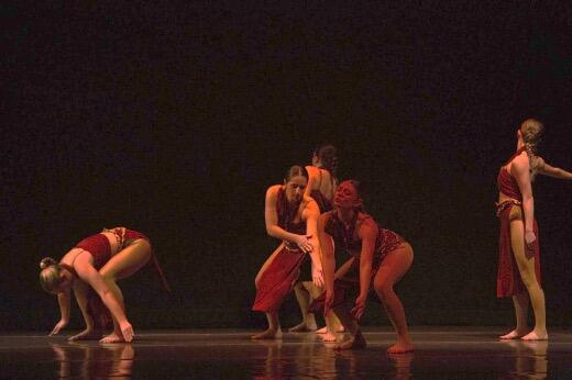 Five dancers in red costumes perform on a dimly lit stage. They are arranged in varied poses, with some bending and others standing. The dark background and reflective stage floor highlight their movements.