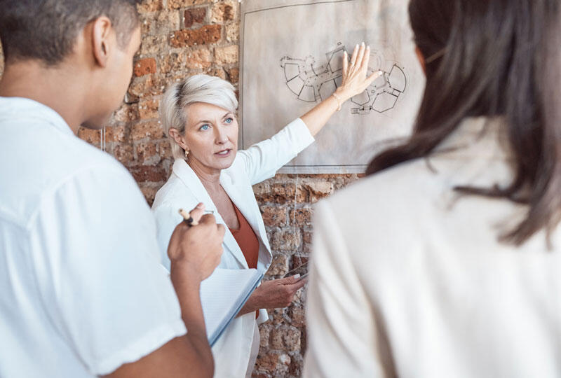 Three people engaged in a discussion, with one person pointing at a technical or architectural diagram on a brick wall.