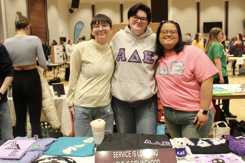 Three Alpha Delta Epsilon members stand behind a table at a social event, displaying items including clothing with Greek letters A delta E and a sign reading 'SERVICE IS LOVE MADE VISIBLE AND LOVE IS OUR SIBLINGHOOD.'