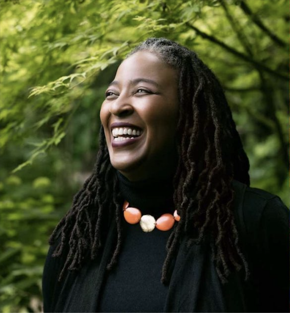 Photograph shows a Black Woman with braids in her hair past her shoulders.  She is wearing a black shirt and black blazer.  She has a big smile on her face, like she is laughing and has a chunky, large beaded necklace on with alternating orange and white beads.  She is in front of lots of greenery, either from trees or bushes.