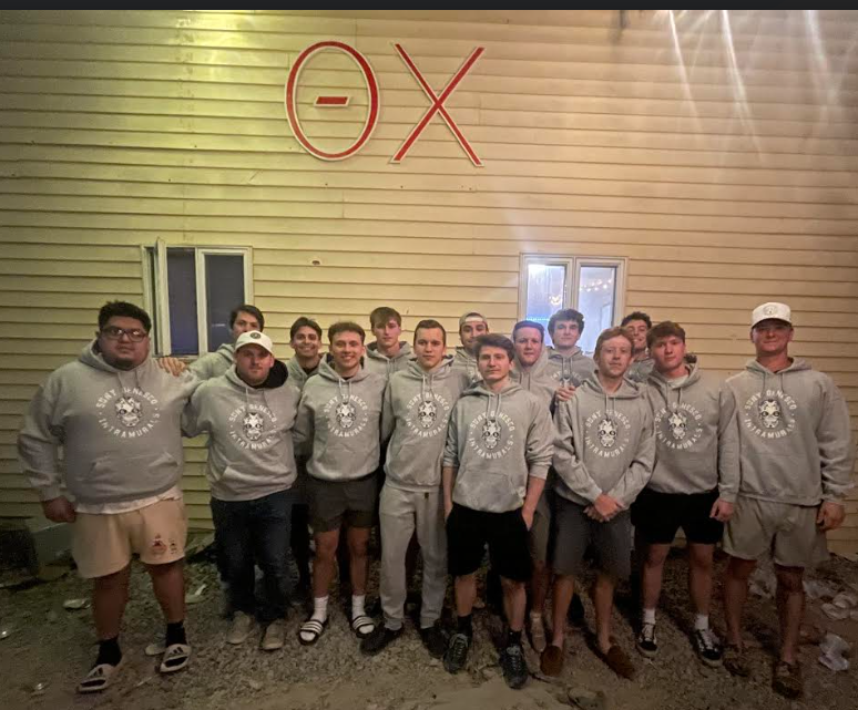 Fifteen athletes wearing gray uniforms and standing together in front of a building