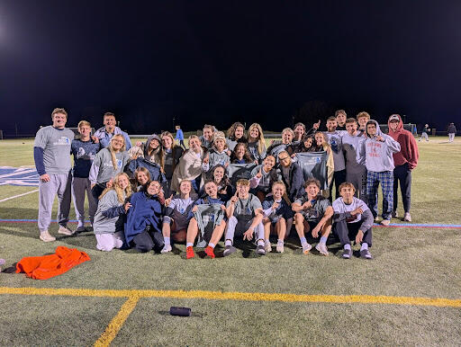 A large group of people on a sports field at night wearing matching gray “SUNY Geneseo Intramurals” shirts, some holding additional shirts, gathered closely for a group photo.