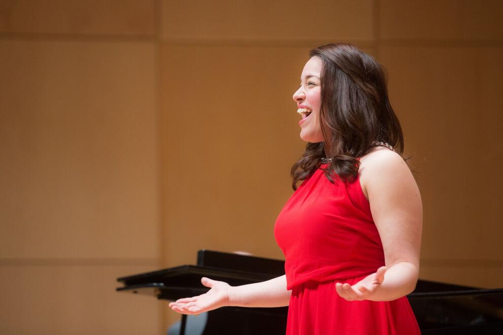 Woman in a red dress standing with arms slightly extended, singing on stage with a piano in the background.