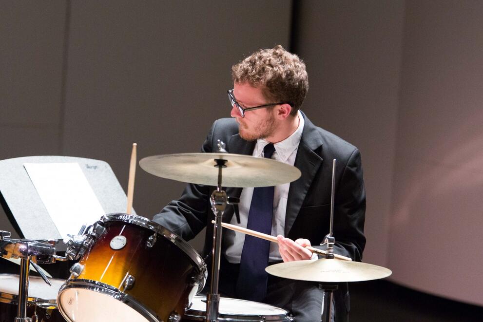 Person in a suit playing a drum set on stage.