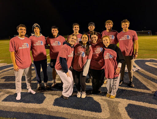 A group of people standing on a sports field at night wearing matching red “SUNY Geneseo Intramurals” T-shirts, posed together on a large logo.