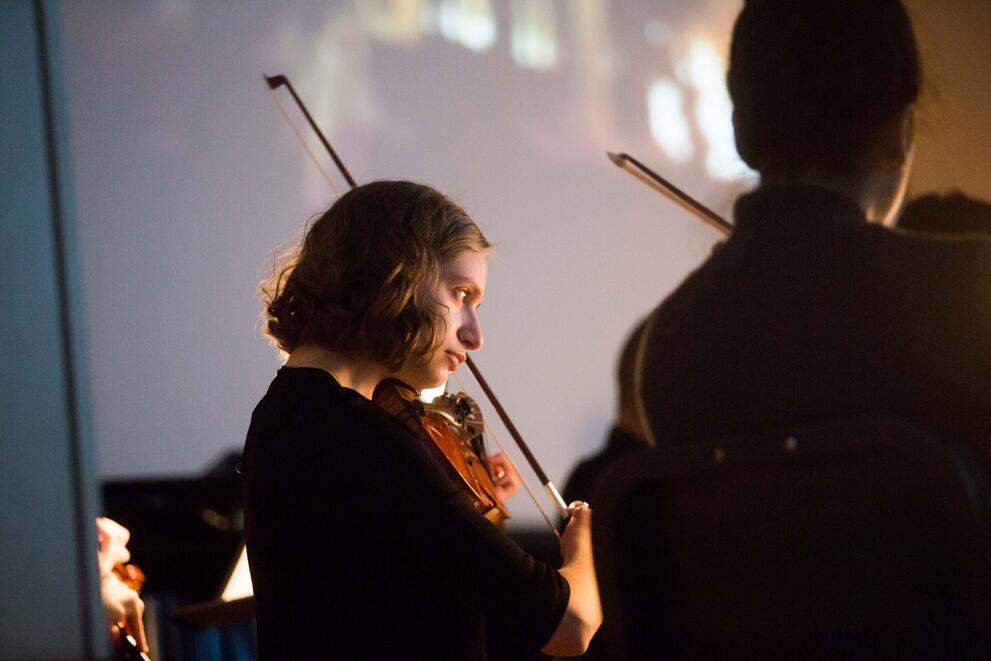 Person playing a violin in an orchestral setting beside another violinist.