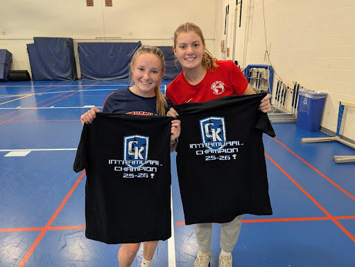 Two students standing on an indoor gym court holding black T-shirts that read “GK Intramural Champion 25-26” in bold blue and white lettering.
