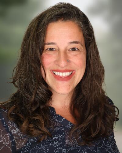 Feminine presenting person with shoulder length brown hair, smiles at the camera.