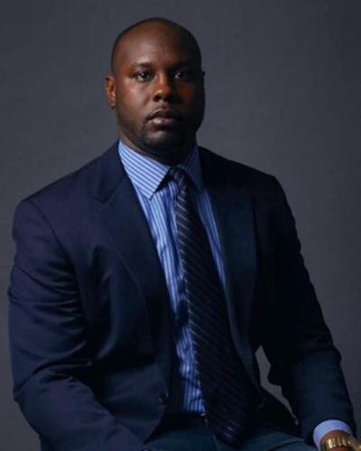 Image of Emmanuel Kulu, Jr. wearing a blue dress shirt and navy suit with a metal watch.  Kulu is seated with his back straight and head facing directly at the viewer.  Kulu looks professional. 