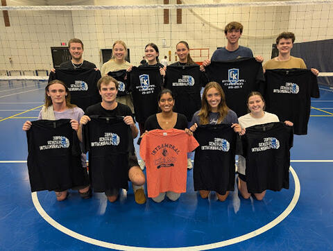 A group of students on an indoor volleyball court holding black “SUNY Geneseo Intramurals” T-shirts, with one person in the front center holding an orange T-shirt.