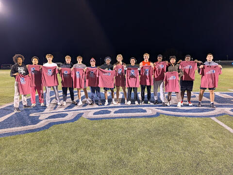 A group of people standing on a sports field at night, each holding a red T-shirt with a logo and text that reads “GK Intramural Champion.”