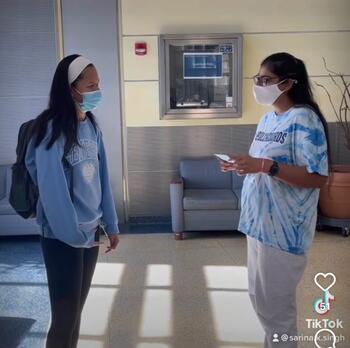 Healthguard Sarina on right wearing a mask and blue tie-dyed shirt talking to a student on left also wearing a mask, blue sweatshirt, white headband, and backpack.