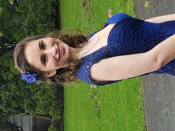 White woman with curled, shoulder length brown hair wearing a blue formal dress and blue flower in her hair