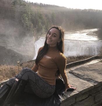 White woman with long brown hair wearing a brown turtle neck sweater sitting on a rock ledge, waterfall behind her