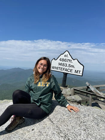 White woman with light brown hair and glasses wearing a dark green jacket sitting on a rock (top of Whiteface Mountain)