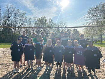 group of students outside on a sand volleyball court holding up grey and black t shirts
