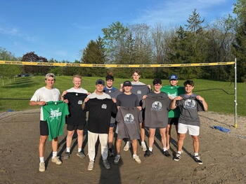 group of students outside on a sand volleyball court holding up grey and black t shirts