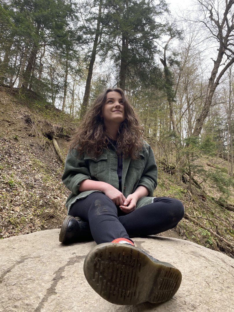 White female with long brown hair sitting on a boulder