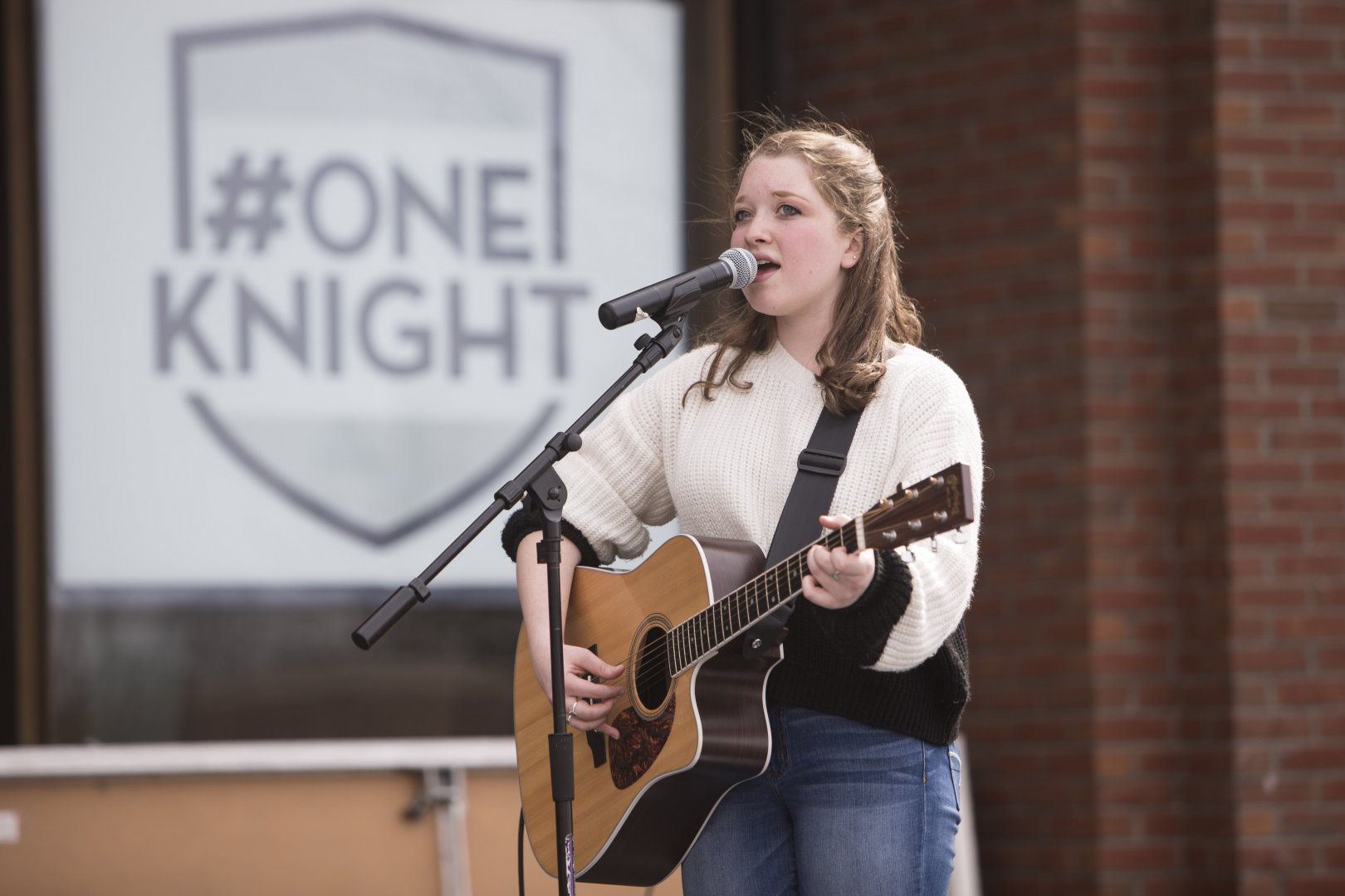 A student, Allison Leah, singing and playing guitar. Behind her, a sign reads hashtag One Knight.