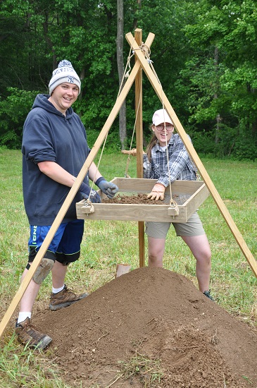 Two people sifting soil through a wooden screen at an outdoor archaeological site. Two people sifting soil through a wooden screen at an outdoor archaeological site.