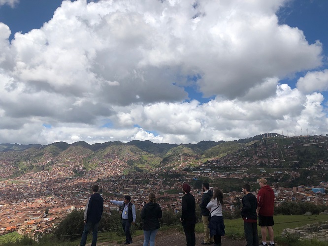 A group of people stands on a hill overlooking a sprawling city in a valley, with a tour guide pointing out something in the distance. The sky is filled with large, white clouds. A group of people stands on a hill overlooking a sprawling city in a valley, with a tour guide pointing out something in the distance. The sky is filled with large, white clouds.
