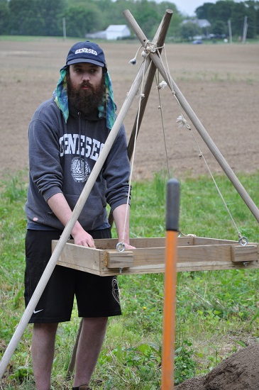 A bearded man uses a screen sifter on a tripod to examine soil at an archaeological dig site in a rural field. A bearded man uses a screen sifter on a tripod to examine soil at an archaeological dig site in a rural field.