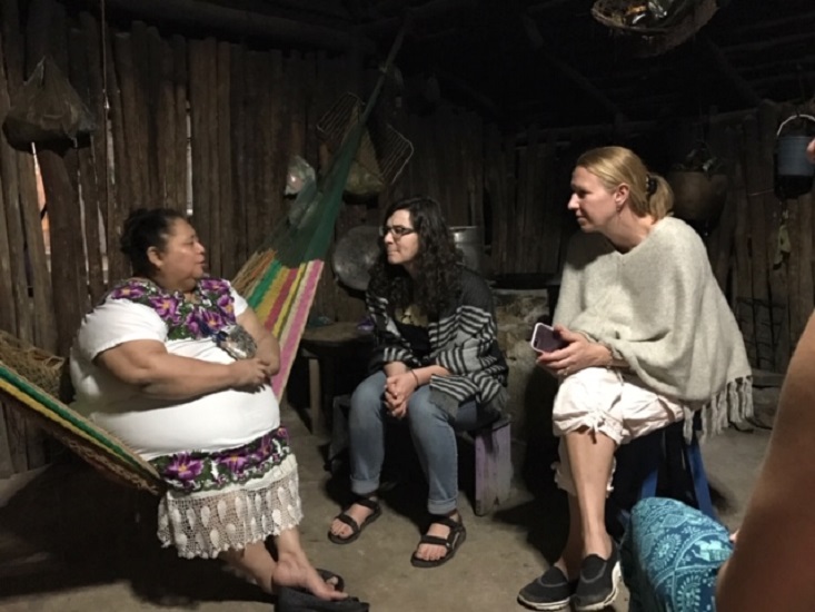 Three women engaged in conversation inside a rustic wooden home, with one sitting in a hammock and the others on stools. Three women engaged in conversation inside a rustic wooden home, with one sitting in a hammock and the others on stools.