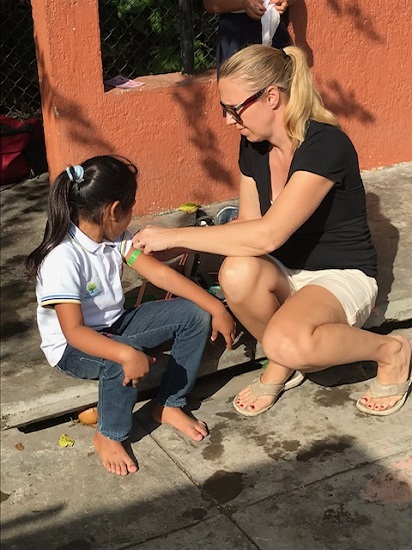 A woman kneels, applying a green bandage to the arm of a young girl with pigtails, who sits outside on a step. A woman kneels, applying a green bandage to the arm of a young girl with pigtails, who sits outside on a step.