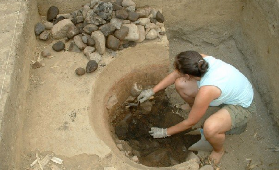 A person is seen in an archaeological dig site, kneeling inside a circular pit filled with soil and debris, while more rocks are piled outside the pit. A person is seen in an archaeological dig site, kneeling inside a circular pit filled with soil and debris, while more rocks are piled outside the pit.
