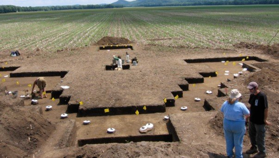 Archaeologists excavate a large dig site in a rural field, uncovering rectangular sections of soil in a cross-shaped layout. Archaeologists excavate a large dig site in a rural field, uncovering rectangular sections of soil in a cross-shaped layout.