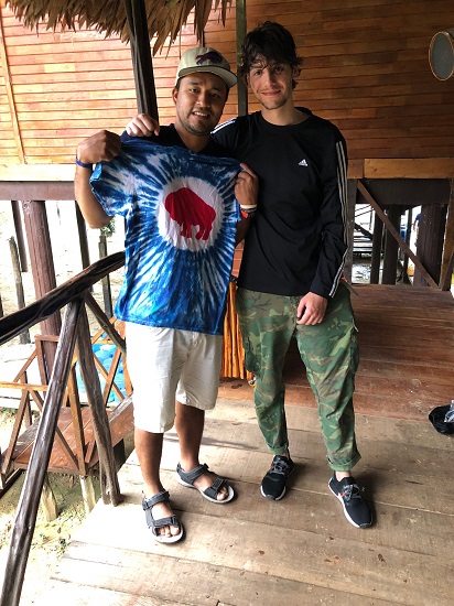 Two men stand smiling on a wooden balcony; one holds up a colorful shirt with a red circle design. Two men stand smiling on a wooden balcony; one holds up a colorful shirt with a red circle design.