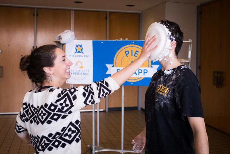 Girl with hand outstretched holding pie in fellow students face.