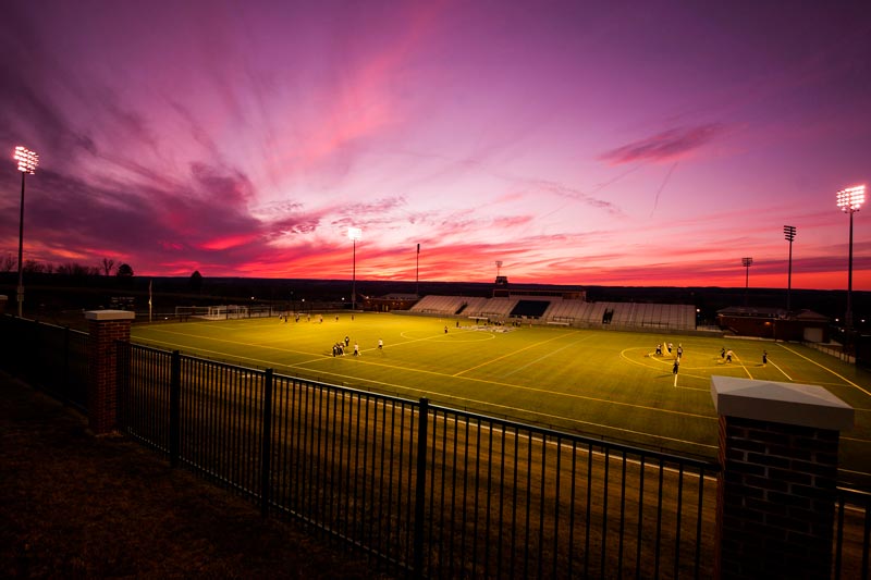 A brightly lit sports field at dusk with a vibrant purple and orange sky, surrounded by fencing and spectators.