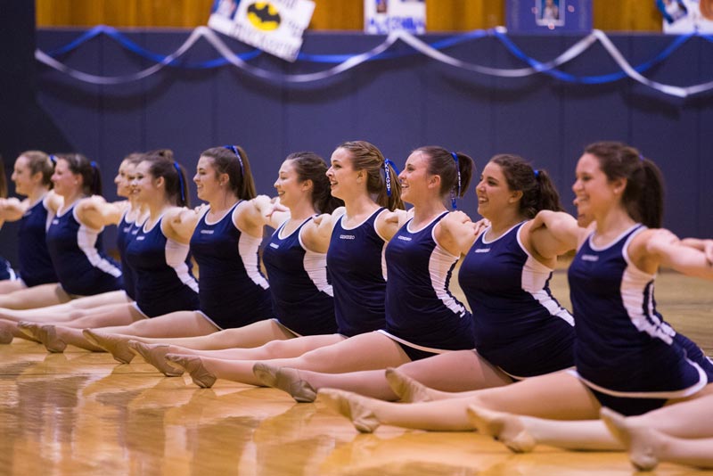 A line of dancers in matching blue and white uniforms sit on a wooden floor, stretching with arms outstretched.