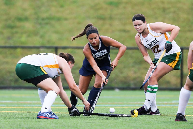 The SUNY Geneseo women's field hockey player about to hit the puck while guarded by two other players The SUNY Geneseo women's field hockey player about to hit the puck while guarded by two other players