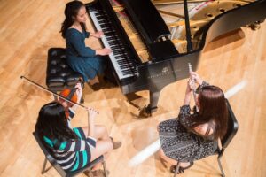 Three musicians perform on a wooden stage: one seated at a grand piano, another playing a violin, and a third playing a flute.
