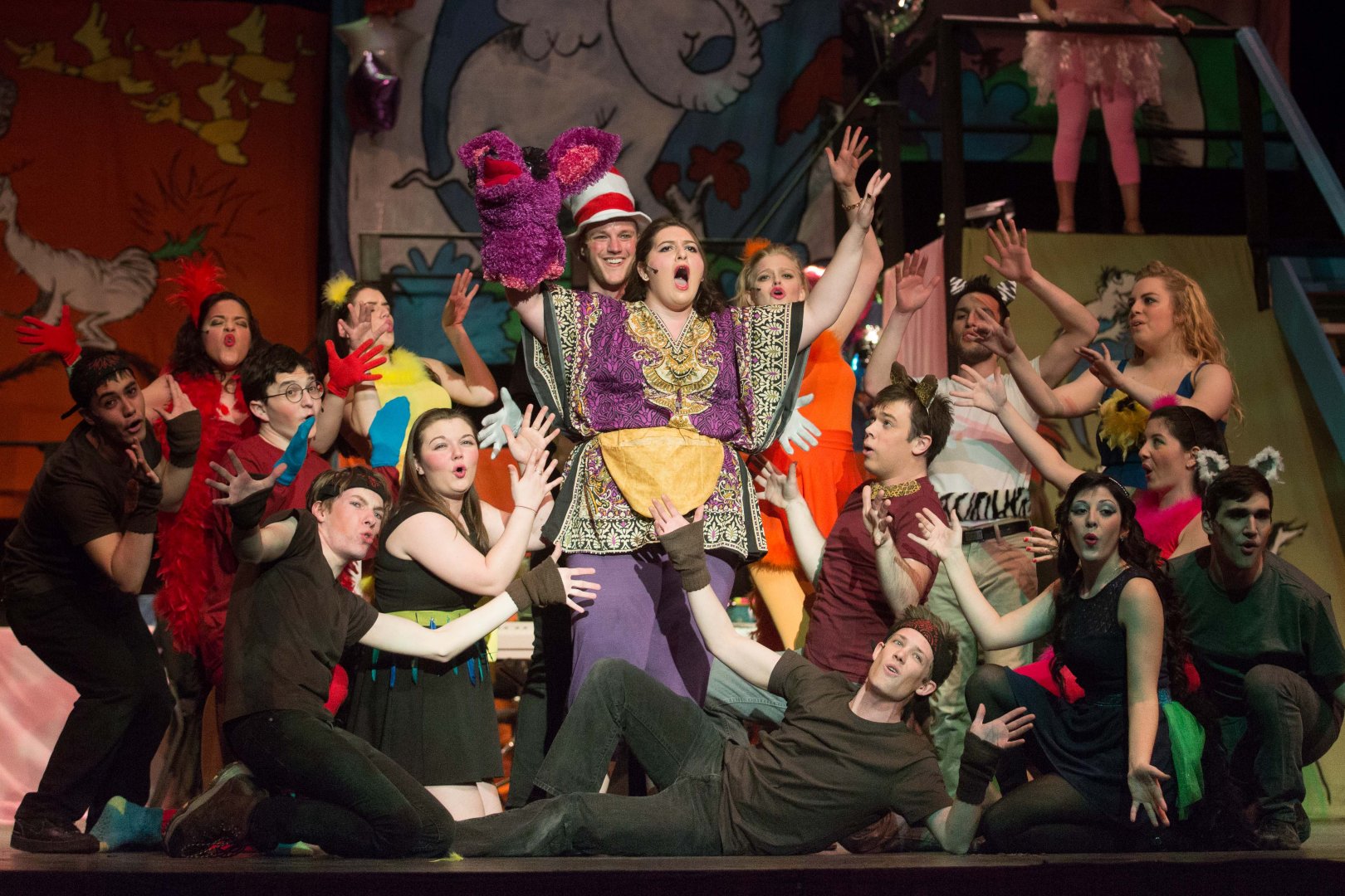 Group of performers on stage in colorful costumes, posing with arms raised or extended. The backdrop features whimsical painted designs, including an elephant and abstract shapes.