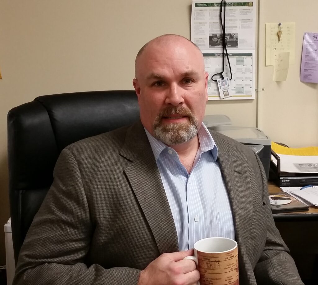 A man sits in a chair facing the camera. He has a goatee and is holding a coffee mug.