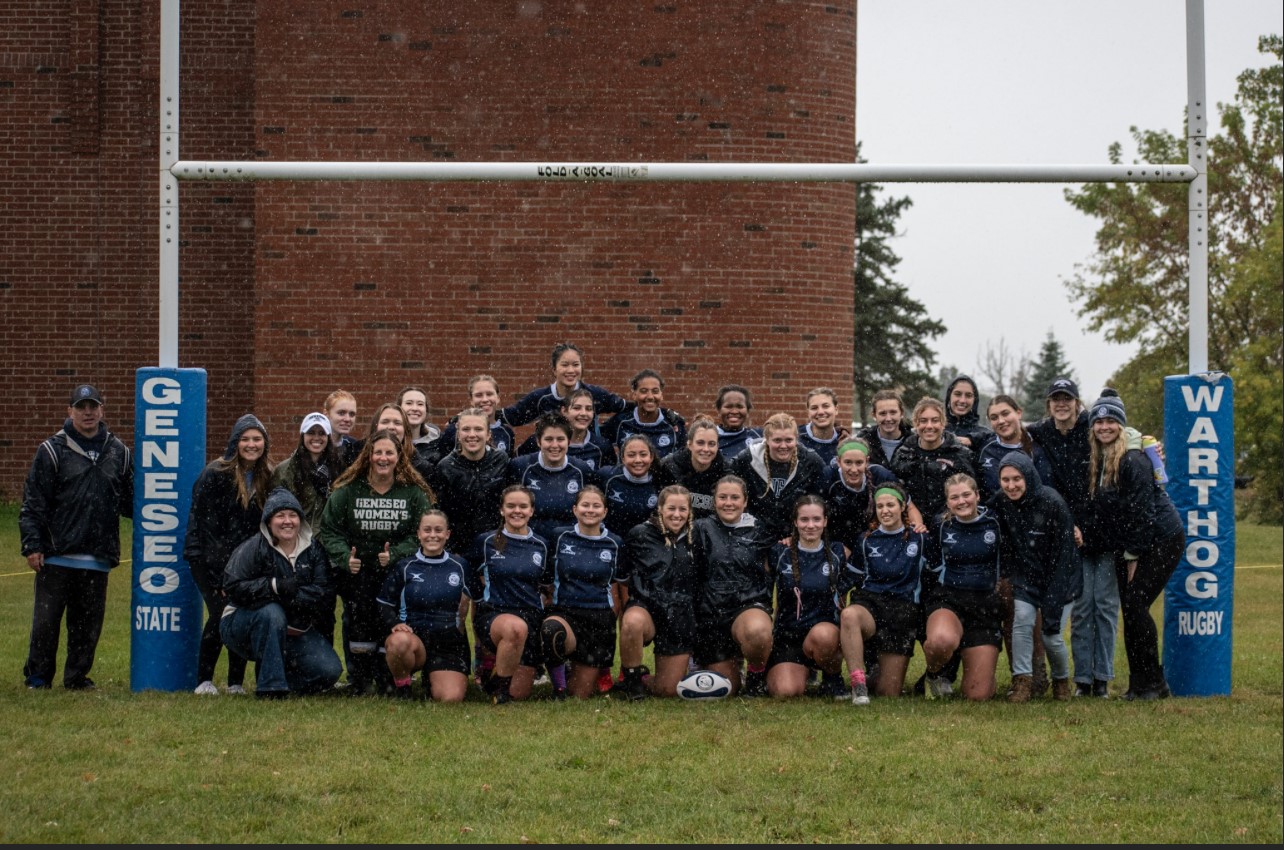 Geneseo Women's Rugby team and their coach posing together next to signs that say Geneseo State Warthog Rugby.