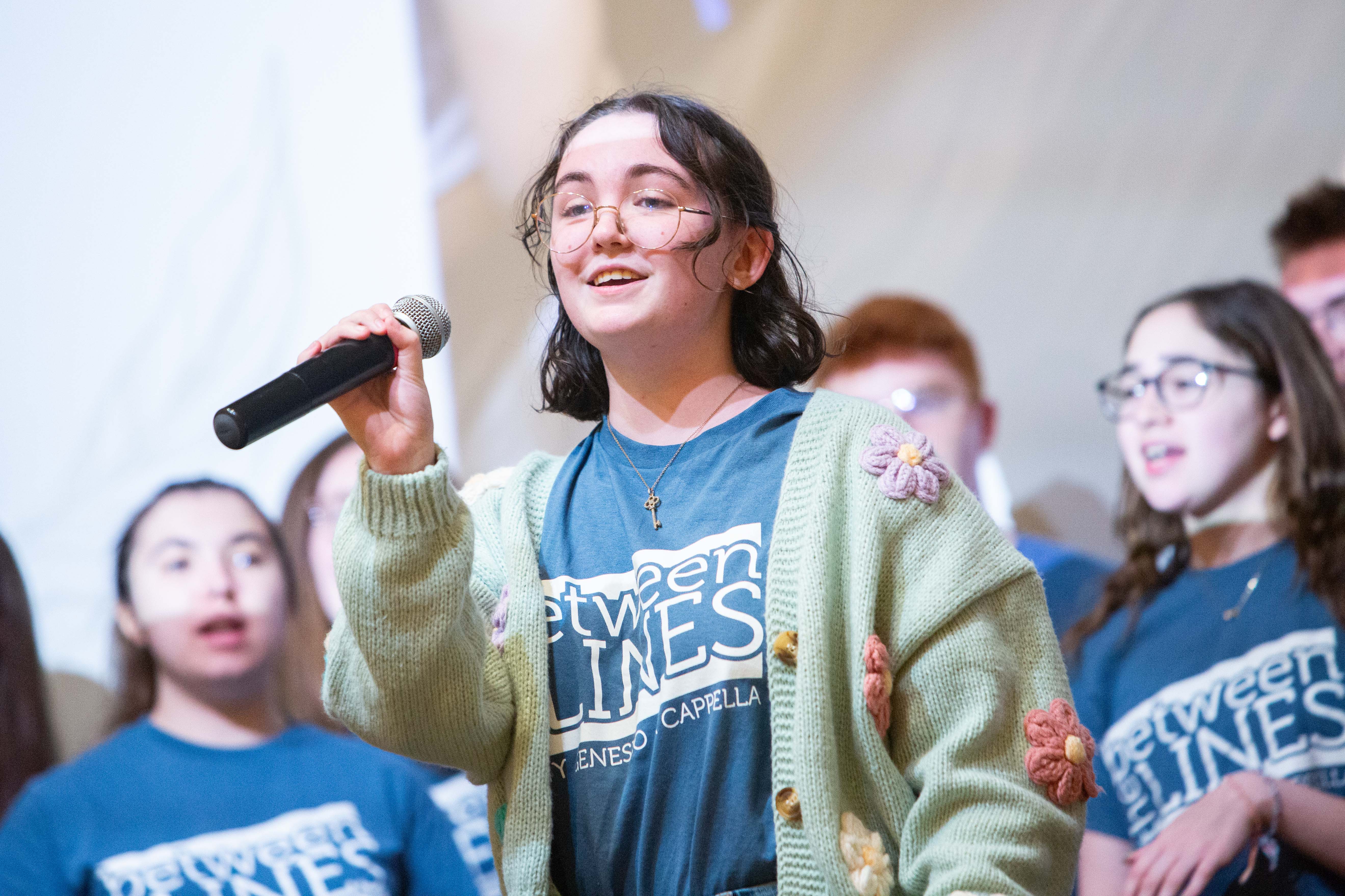 Student wearing a Between Lines Geneseo Acapella shirt and speaking into a microphone.