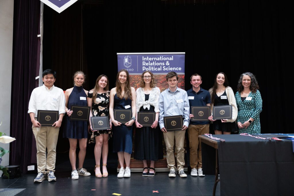 Eight students and a professor stand on a stage holding their awards certificates.