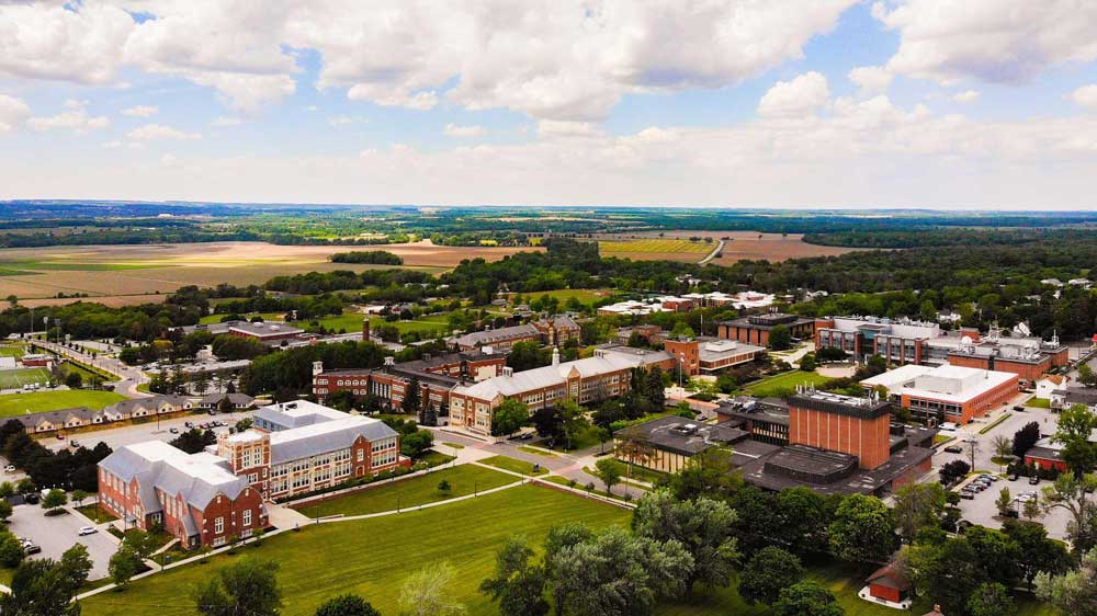 geneseo-campus-aerial-view_0 Aerial view of SUNY Geneseo campus with multiple brick and modern buildings, green lawns, trees, and surrounding farmland under a partly cloudy sky.