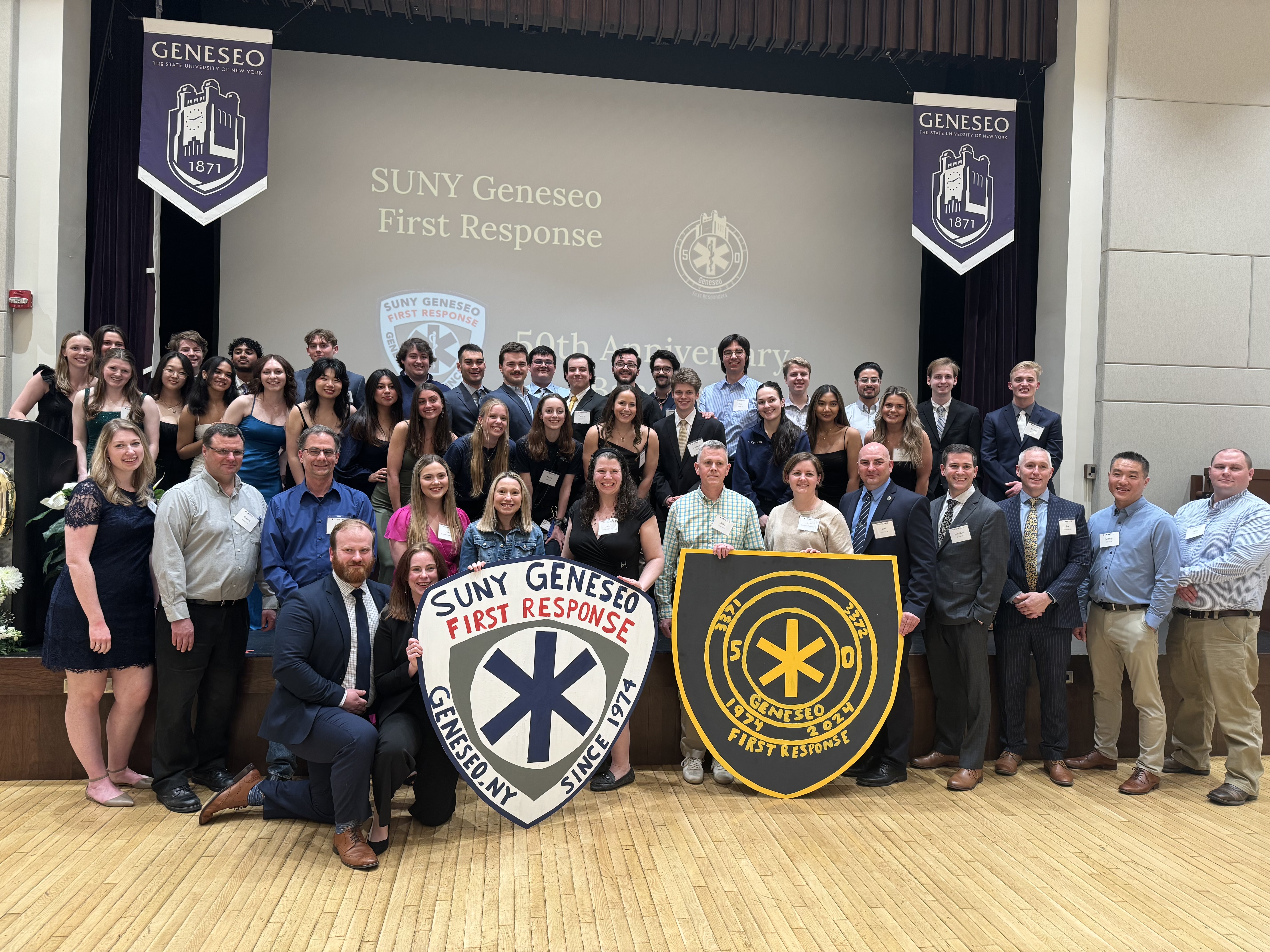 Large group of people standing on a stage in front of a slide that says SUNY Geneseo First Response 50th Anniversary