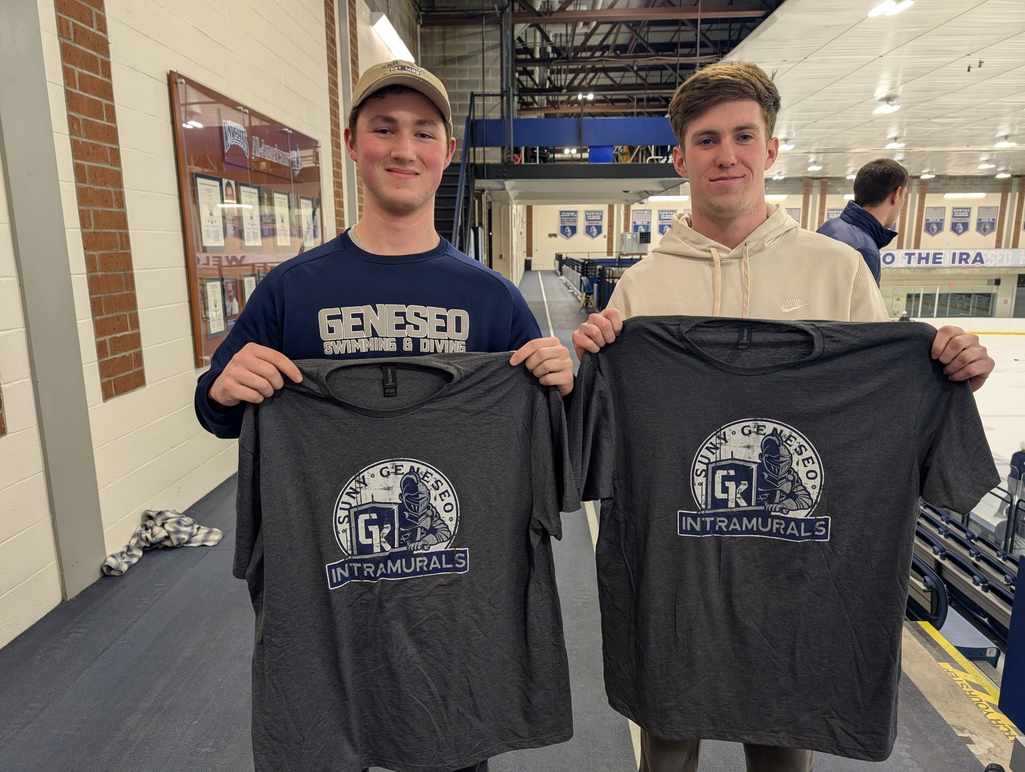 alt="2 students, holding up grey intramural shirts, on the track above ice rink"